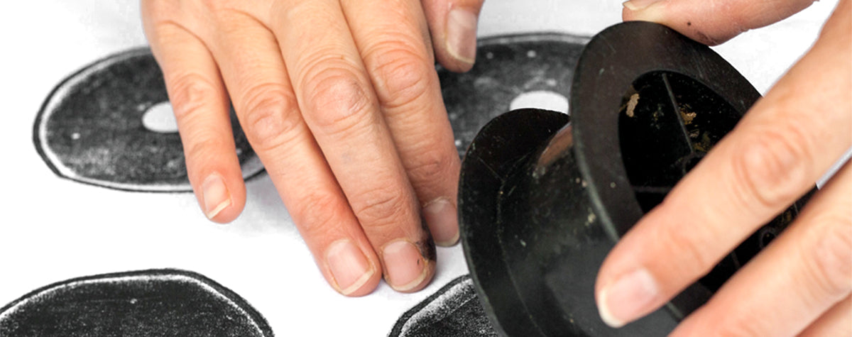 A pair of white hands are block printing, black circles onto white reclaimed cotton fabric, using a discarded black fishing reel, one hand is lifting up the reel, to reveal the print and the other is gently holding down the reclaimed cotton fabric.