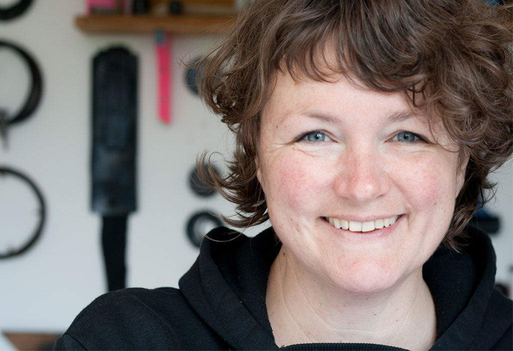 A white female printmaker faces the camera and smiles. In the background are found beach items hanging against white wooden walls of a studio interior.