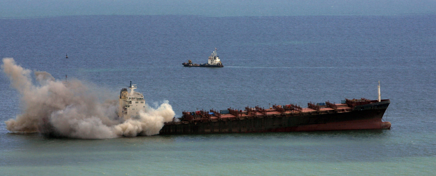 An empty MSC Napoli, floats in calm water. Vast plumes of smoke are billowing from around the bridge of the vessel. In the distance a tug boat can be seen.