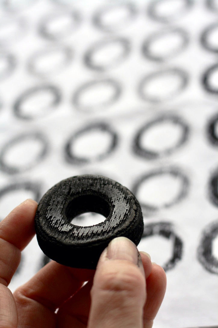 A hand holds up a found piece of circular black rubber. Behind, a piece of white cotton fabric, is printed with black circles.