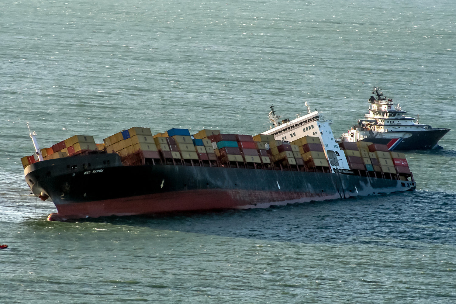 The MSC Napoli, a 900ft long cargo ship, lists heavily away from the camera. It is carrying 2,300 containers. To the rear of the ship, there is a rescue vessel.