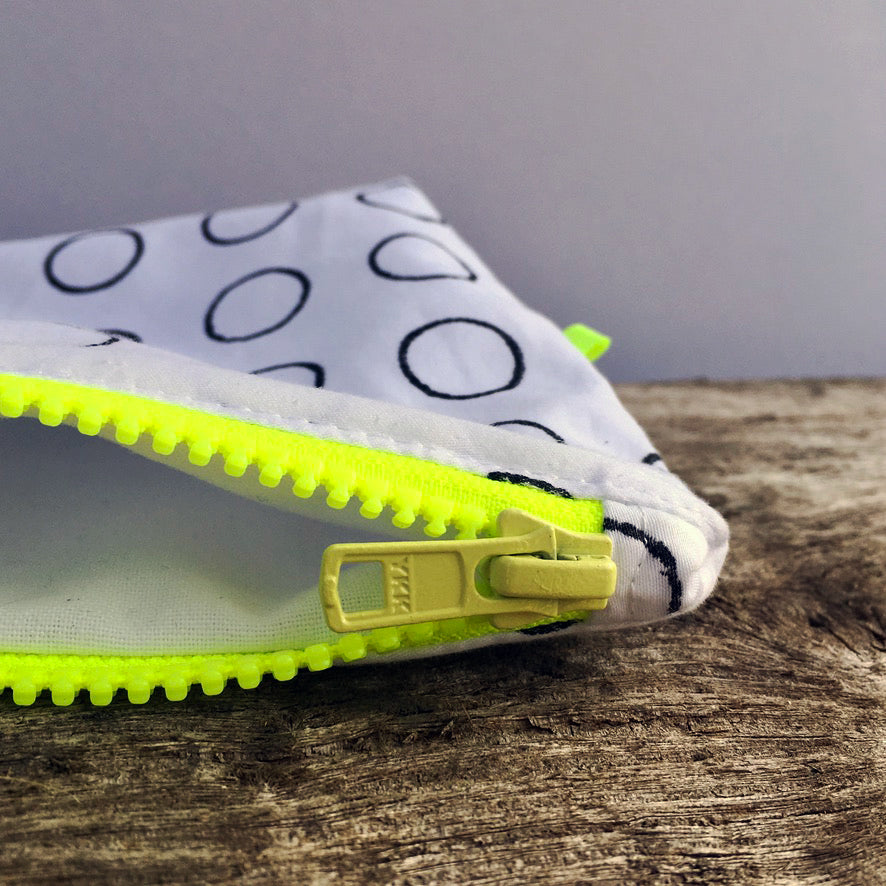 Close up of handmade white reclaimed cotton purse, with black rings printed on it. The YKK chunky neon yellow zip, is the focus in the foreground, and it is sat on a piece of weathered driftwood, against a grey background.