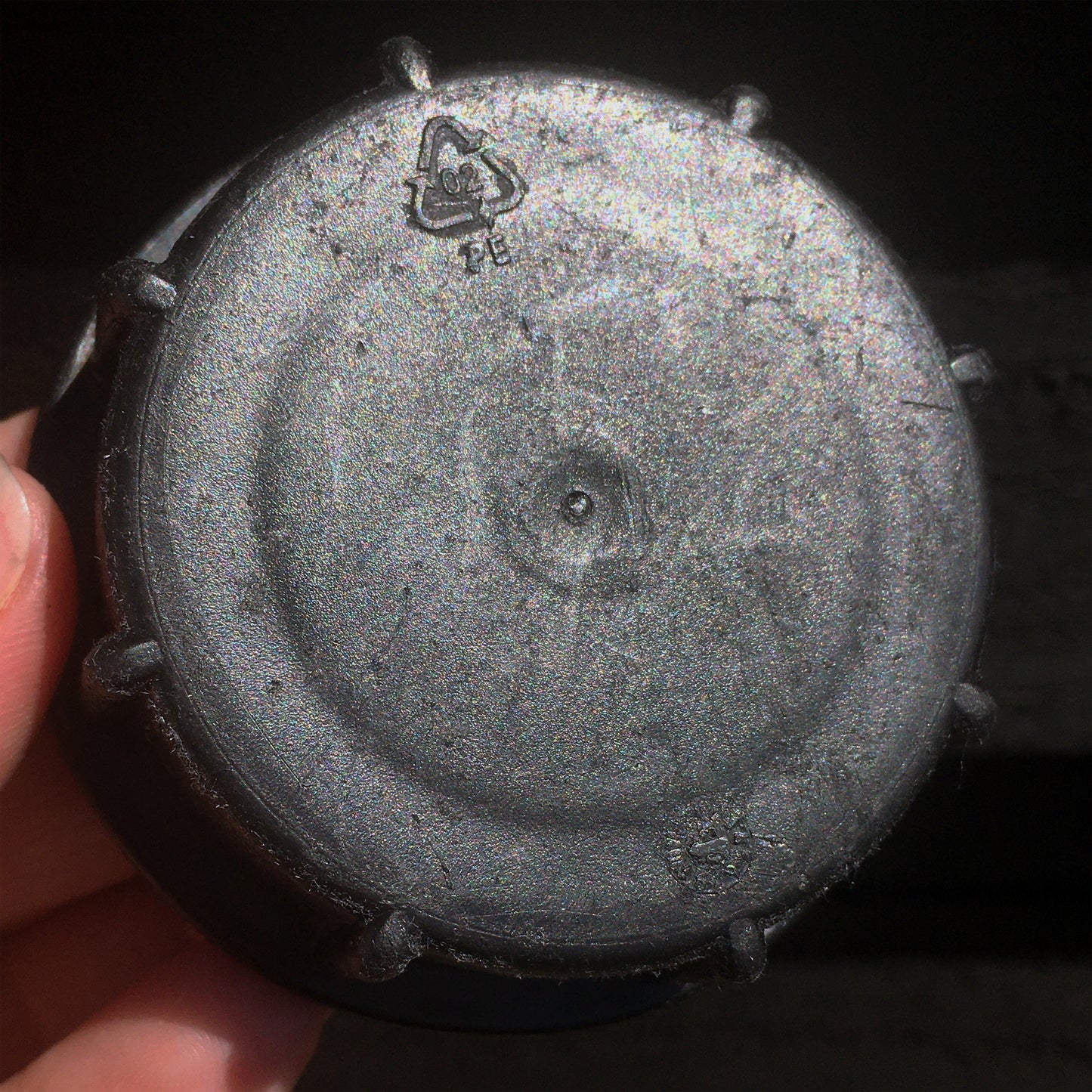 A white hand holds a discarded black plastic drum cap. It features a recycle symbol with the number 02, in it.
