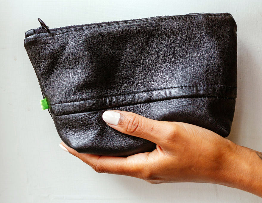 A black woman's hand is holding a soft black leather purse, against a white wall.