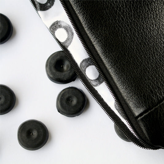 A close up image of a soft, black leather purse with hand printed lining. The lining is white cotton with small black rings on it. The purse is lying against a white surface. To the left of the image, are four small black rubber blanking plugs.
