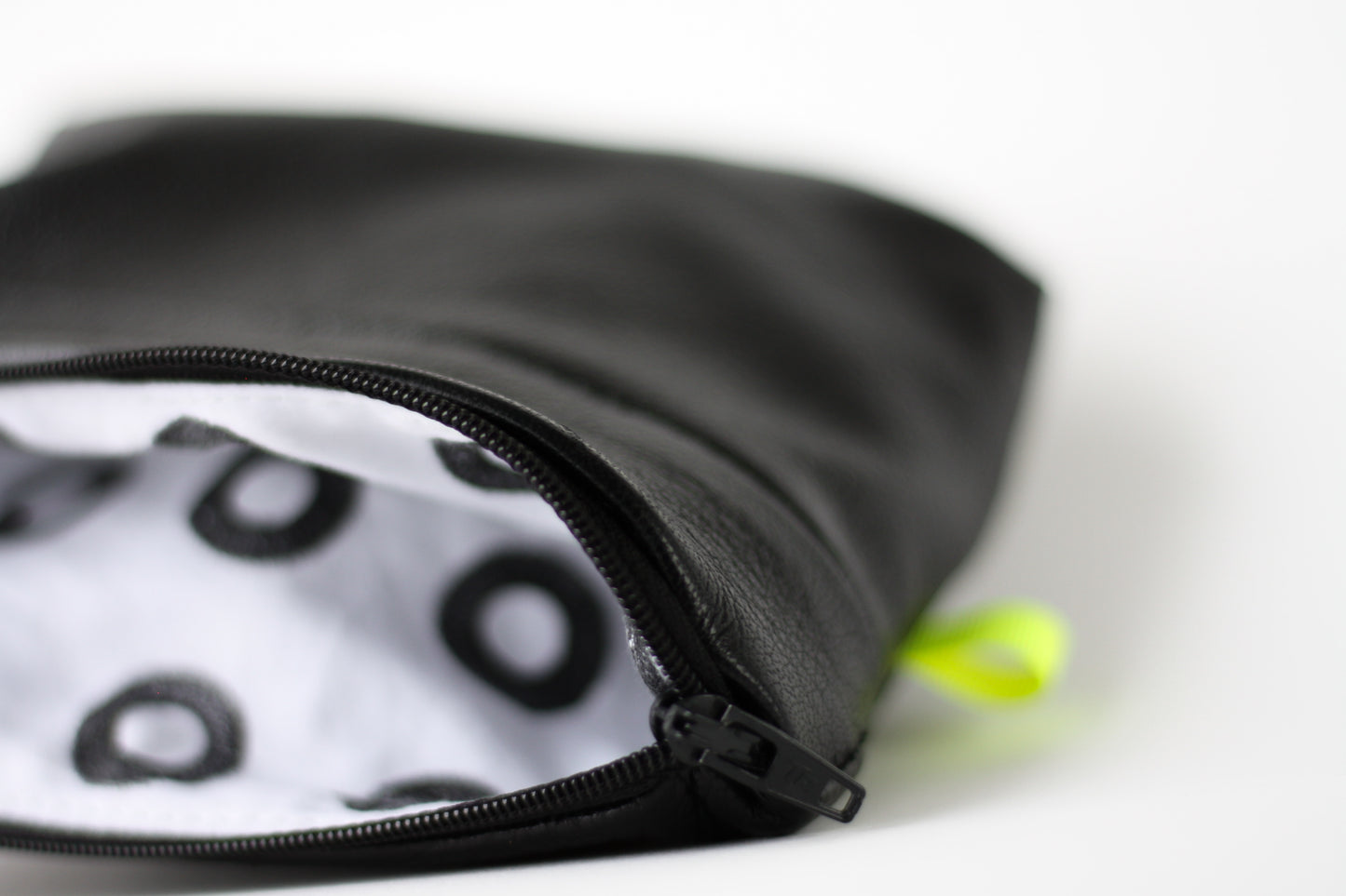 A handmade black leather purse, lies on its side, to reveal a hand printed lining. The lining is white cotton with black rings. The purse has a small neon yellow tab sewn into the side seam.