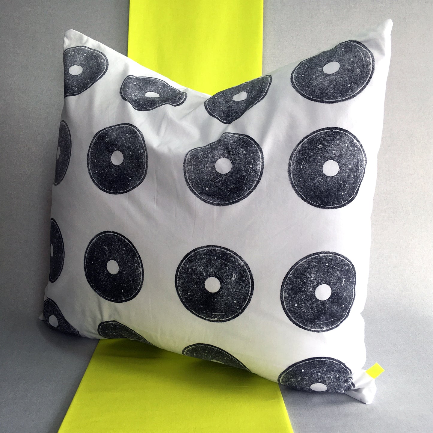 A sustainable hand printed cushion. Made from white reclaimed cotton with hand printed black circles, made using a discarded fishing reel. The cushion sits on a strip of neon yellow fabric against a grey background.