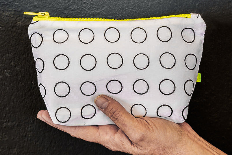 A white reclaimed cotton purse with hand printed black rings, is sat in a white woman's hand, against a black painted wall. It features a chunky YKK neon yellow zip, and neon yellow recycled polyester tab.