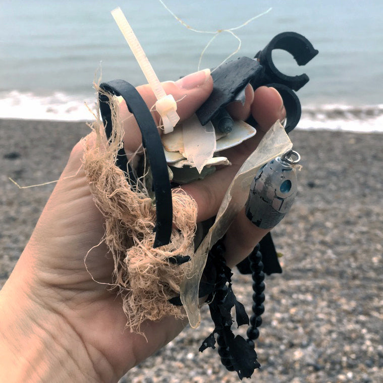 A white hand holds up found items from a beach clean. Pebbles and sea are in the background.