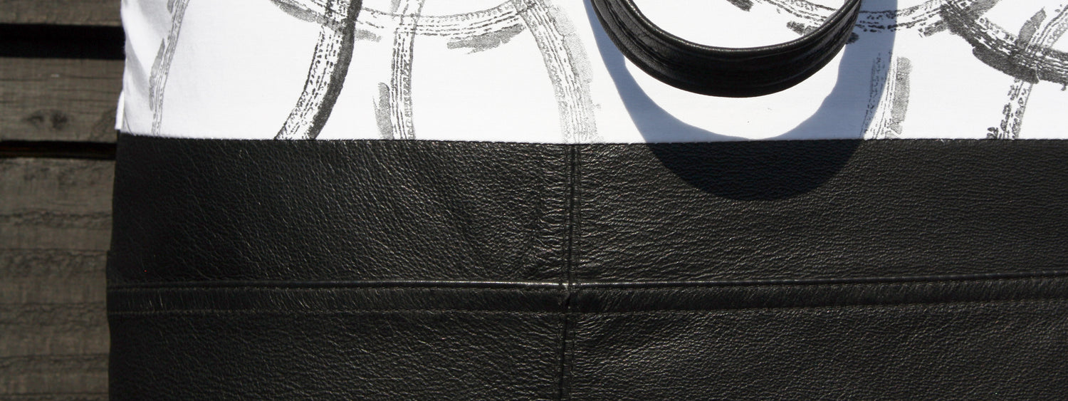 A close up image of a black and white leather and hand printed cotton bag. The white cotton has overlapping textured, black rings on it. It reads Handmade Bags over the top of the black leather. And underneath it, One of a kind bags with a story.