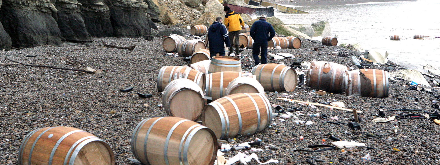 Oak barrels lie damaged on a pebble beach, as three men walk amongst them and other debris, washed ashore from the MSC Napoli. In the distance shipping containers can be seen.