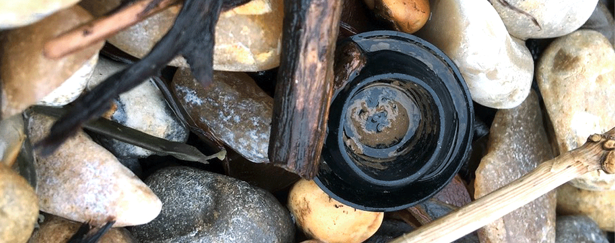 A close up of a small black rubber blanking plug, sits upturned, amongst pebbles and driftwood sticks. The words Create From Waste, collected from our beautiful Devon coast, is written across the image.