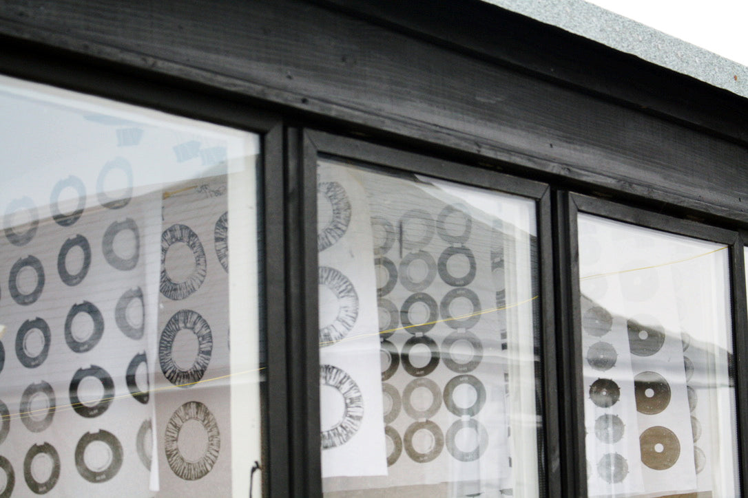Looking through windows of a black painted, wooden studio. Hand printed white cotton, fabrics hang, with black printed circles on them.