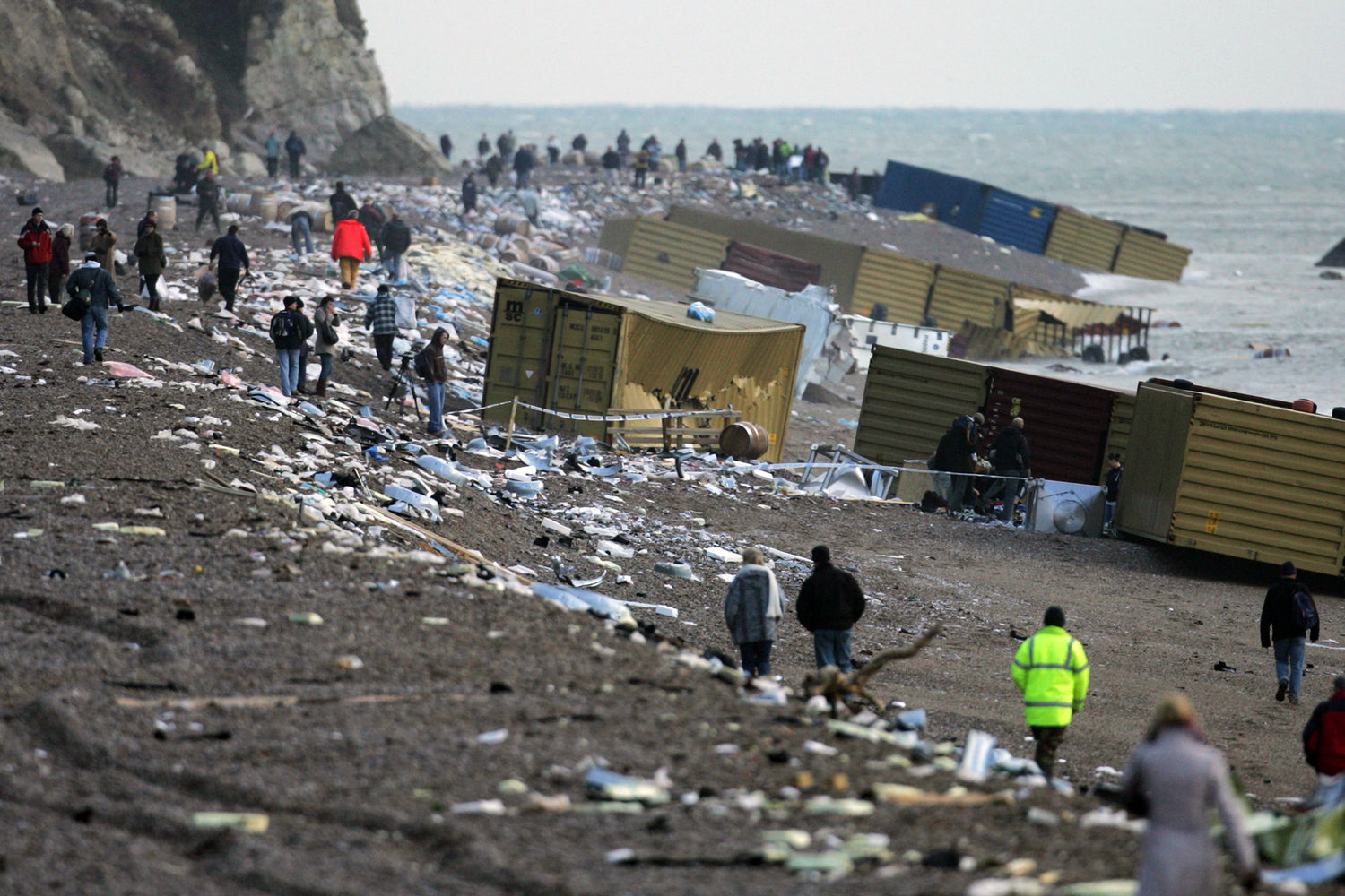 Twelve containers from the MSC Napoli cargo spill, can be seen lying on their sides on a pebble beach. Some have torn open. The contents of the containers are strew all along the length of the beach. People wearing winter clothes, walk amongst the debris.