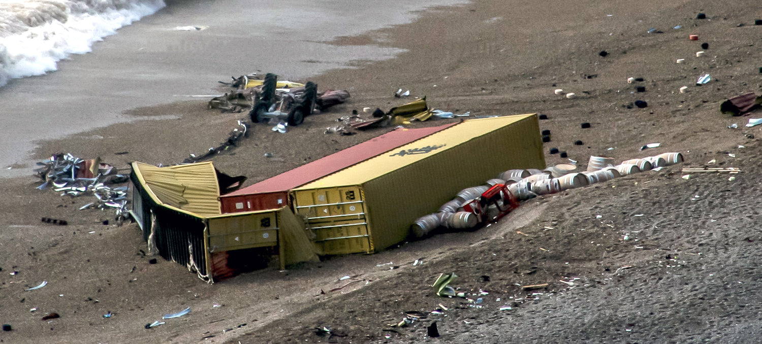 Three shipping containers are lying on their sides, one is crumpled open. Next to them lie oak barrels and large pieces of metal. In the distance an upturned tractor chassis, is lying at the tideline. 