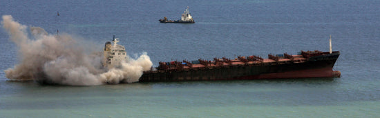An empty MSC Napoli, floats in calm water. Vast plumes of smoke are billowing from around the bridge of the vessel. In the distance a tug boat can be seen.