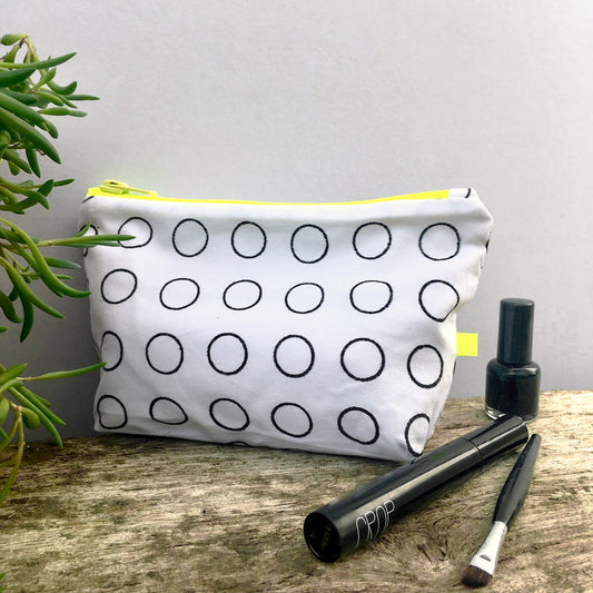 A white reclaimed cotton purse sits on a piece of driftwood. It has a neon yellow zip, tab and black rings printed on it. It sits next to make up and a succulent plant.