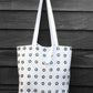 A black and white hand printed tote bag, hangs against a black painted wooden surface.The bag features a grid of small black printed rings and dual handles.