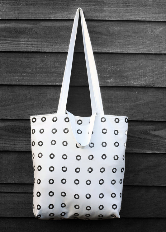 A black and white hand printed tote bag, hangs against a black painted wooden surface.The bag features a grid of small black printed rings and dual handles.