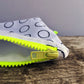 Close up of handmade white reclaimed cotton purse, with black rings printed on it. The YKK chunky neon yellow zip, is the focus in the foreground, and it is sat on a piece of weathered driftwood, against a grey background.