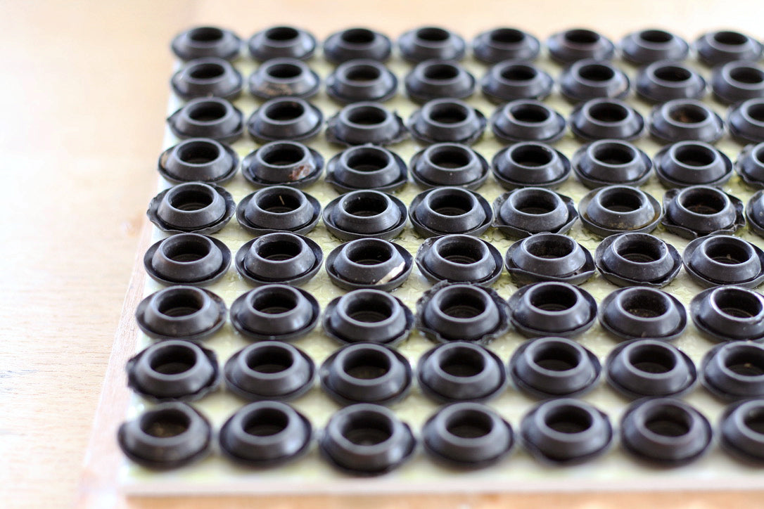 A series of 81 upturned rubber blanking plugs, form a grid. They are glued to a piece of reclaimed wooden board. The board sits on a wooden table.