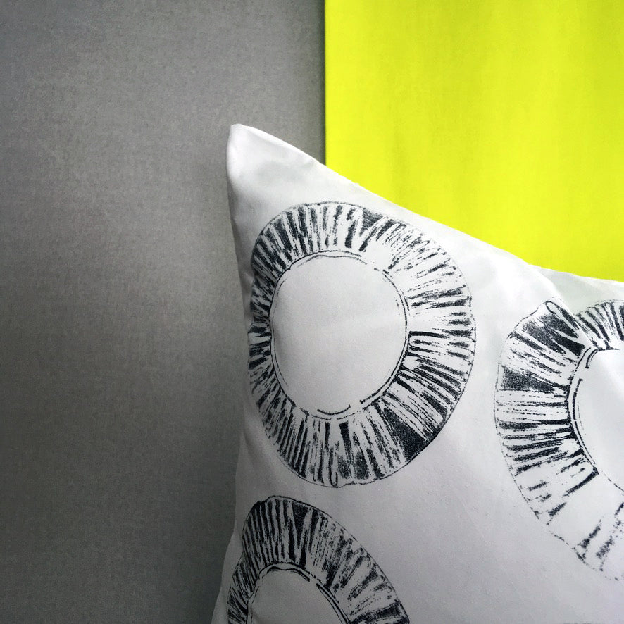A close up detail, of the corner of a hand printed cushion. The print is a black textured ring, created using a car filter. The cushion sits against a strip of yellow fabric and a grey background.
