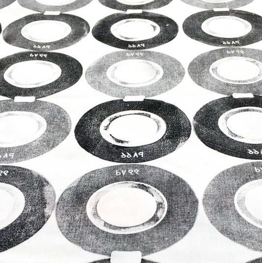 Close up of hand printed fabric, featuring a repeat pattern of circular discs, in varying tones of black and grey. Each discs has PA66 written in reverse.