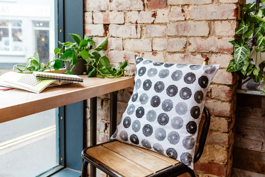 A black and white, sustainable hand printed cushion sits on a metal and wooden, industrial style stool, against a red brick wall. The print is a made from a discarded plastic drum cap, in varying shades of black and grey.