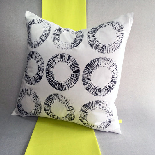 A black and white hand printed cushion sits on a strip of neon yellow fabric, against a grey background. The cushion features 9 circular prints, in varying shades of black, made from a found car filter.