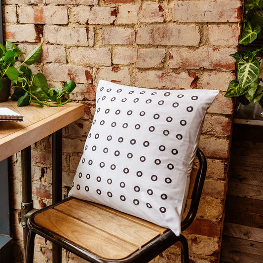 A black and white hand printed cushion, sits on an industrial style, wooden and metal stool. Made from sustainable, reclaimed cotton, it features a grid of black rings.