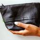 A black woman's hand is holding a soft black leather purse, against a white wall. 