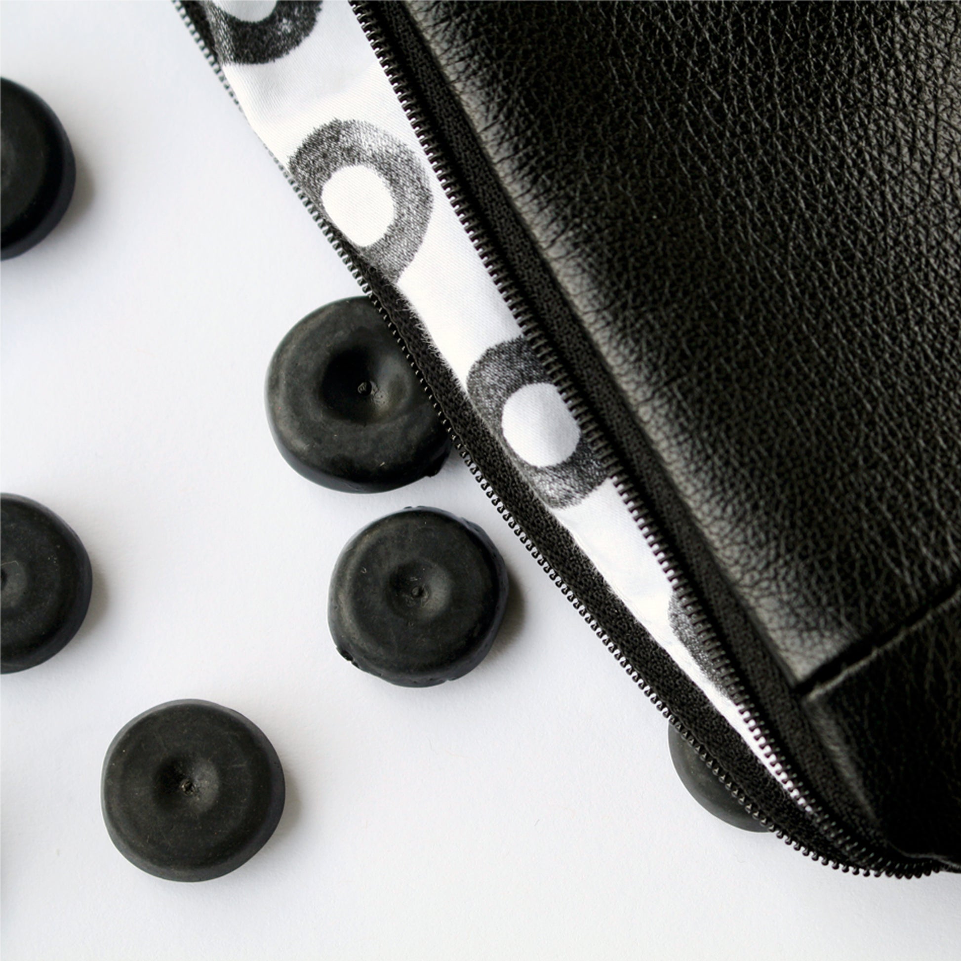 A close up image of a soft, black leather purse with hand printed lining. The lining is white cotton with small black rings on it. The purse is lying against a white surface. To the left of the image, are four small black rubber blanking plugs. 