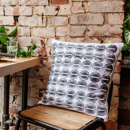 A hand printed sustainable cushion sits on a wood and metal industrial style stool, against a red brick wall. The cushion features a black circular block print on white reclaimed cotton. 