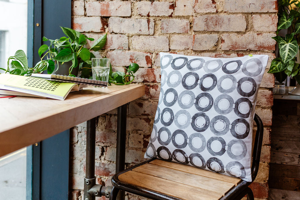 A sustainable hand printed cushion, featuring block printed rings, sits on a wood and metal, industrial style stool, against a red brick wall. There are trailing plants and books in the window.