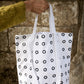 A white woman's hand holds up a white cotton tote bag, with small black hand printed rings on it, against a background of monastic walling. 