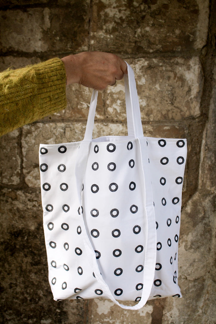 A white woman's hand holds up a white cotton tote bag, with small black hand printed rings on it, against a background of monastic walling. 