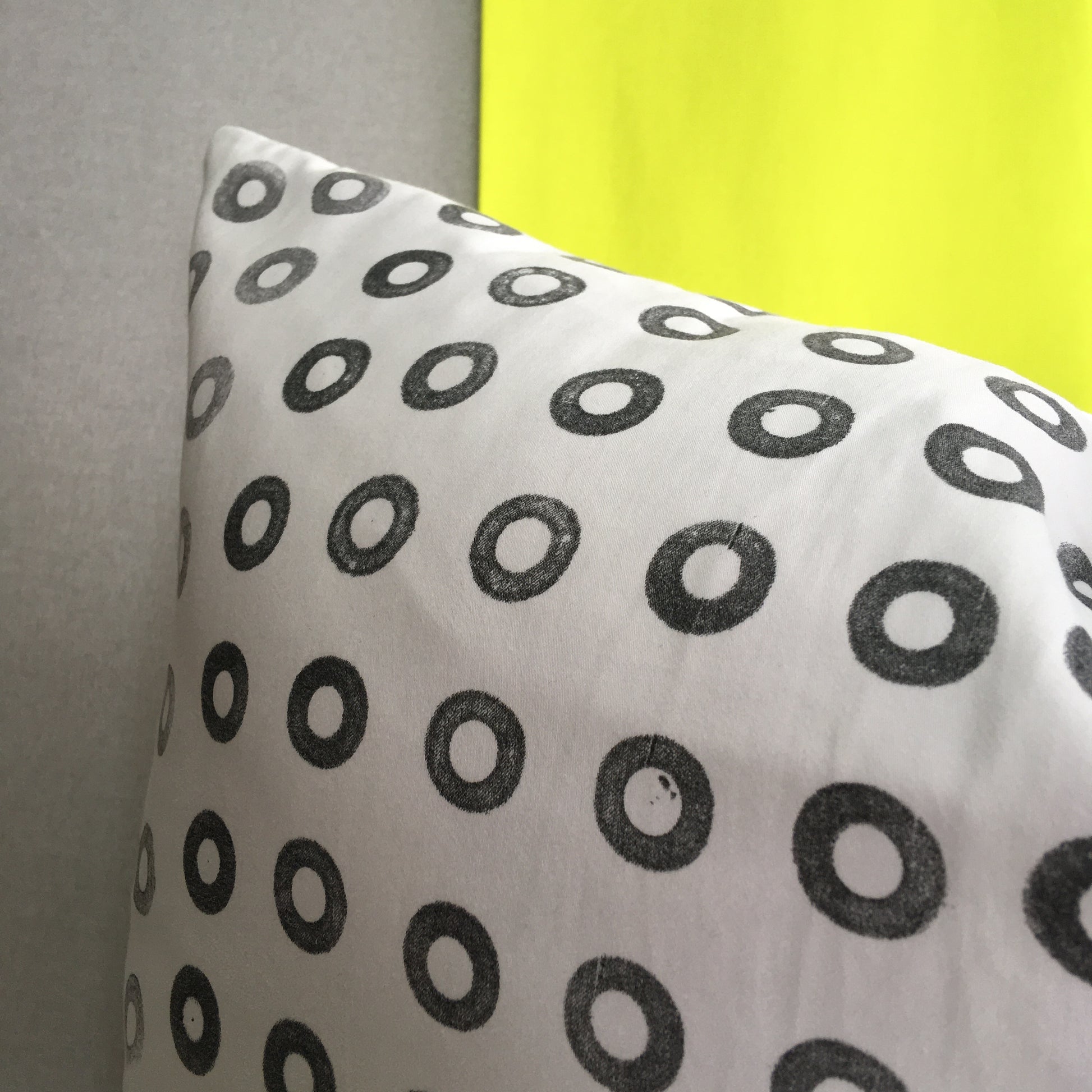 A hand printed cushion, made from reclaimed cotton, sits against a grey and acid yellow background. It features a grid of small black rings, printed using found beach waste.