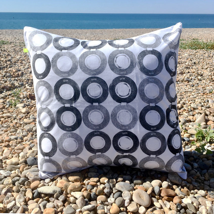 A hand printed black and white cushion, sits on a pebble beach, with the sea in the background. 