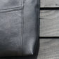  A close up detail of a soft black leather bag, against a black painted wooden surface. The image focusses on the original stitching on the leather.
