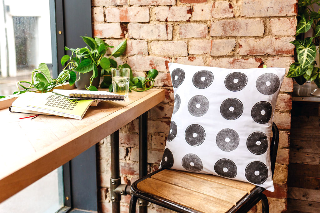 White reclaimed cushion with black printed circles, made from a discarded fishing reel. The cushion is sitting on a wooden and metal industrial style stool, against a red brick wall. There are plants and books in the window.