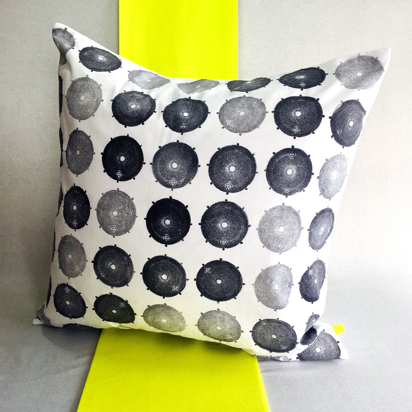 A hand printed black and white sustainable cushion, sits on a strip of neon yellow fabric, against a grey background. The print features a drum cap in varying tones of black and grey.