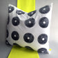 A sustainable hand printed cushion. Made from white reclaimed cotton with hand printed black circles, made using a discarded fishing reel. The cushion sits on a strip of neon yellow fabric against a grey background.