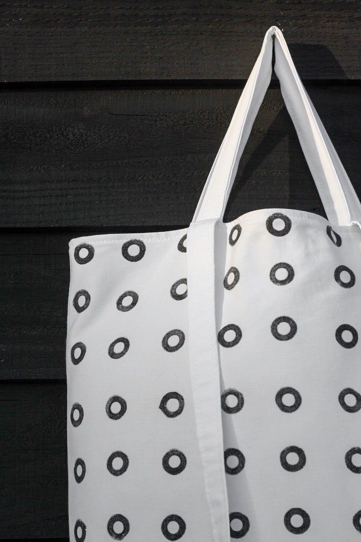 A close up of a black and white hand printed cotton tote bag, featuring small black rings. The bag hangs against a black painted wooden surface.