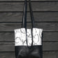 A black and white hand made tote bag hangs against a black wooden surface. It has black leather straps and soft leather base, made from reclaimed leather jackets. The top half of the bag, features hand printed overlapping circles in varying shades of grey and black, printed onto white cotton, using a piece of found rubber, from a beach clean.