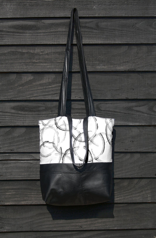 A black and white hand made tote bag hangs against a black wooden surface. It has black leather straps and soft leather base, made from reclaimed leather jackets. The top half of the bag, features hand printed overlapping circles in varying shades of grey and black, printed onto white cotton, using a piece of found rubber, from a beach clean.