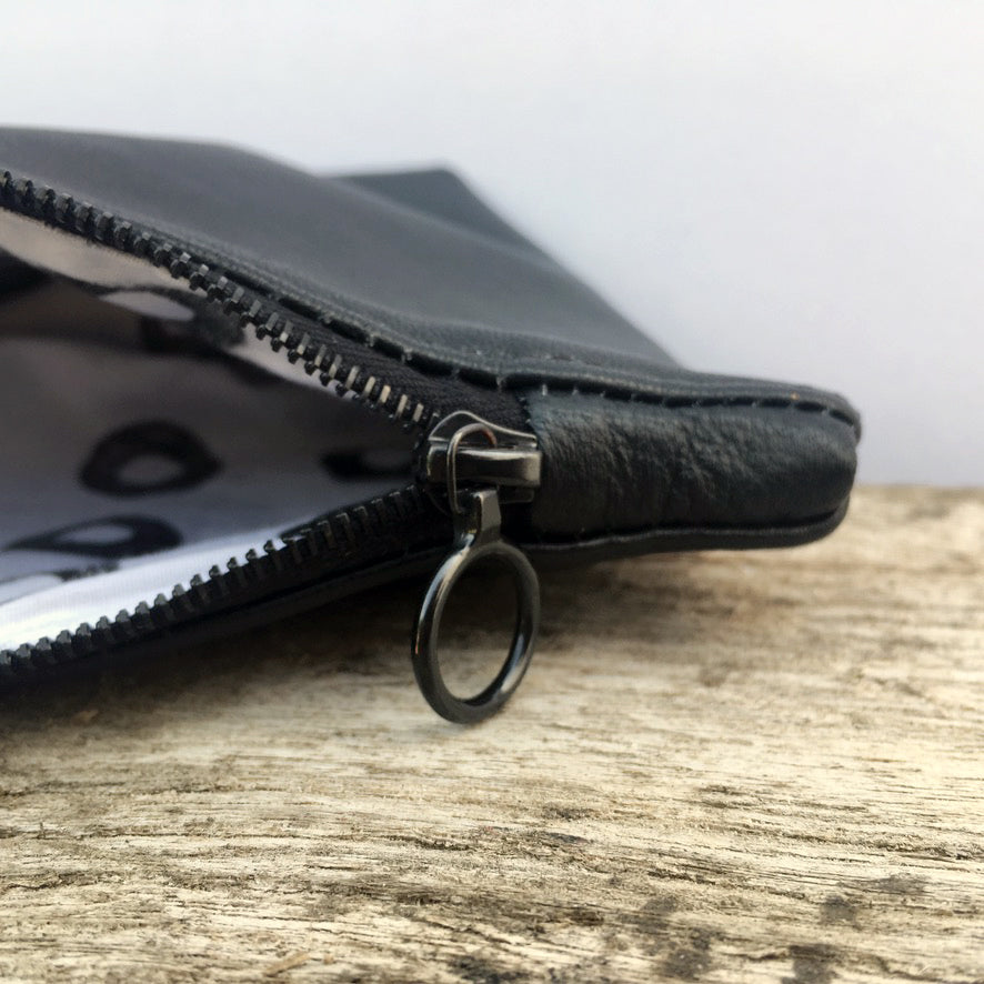 A close up image of a black soft leather purse. The image focusses on a black circular recycled Riri zip. The purse is lying on a piece of driftwood, against a grey background. 