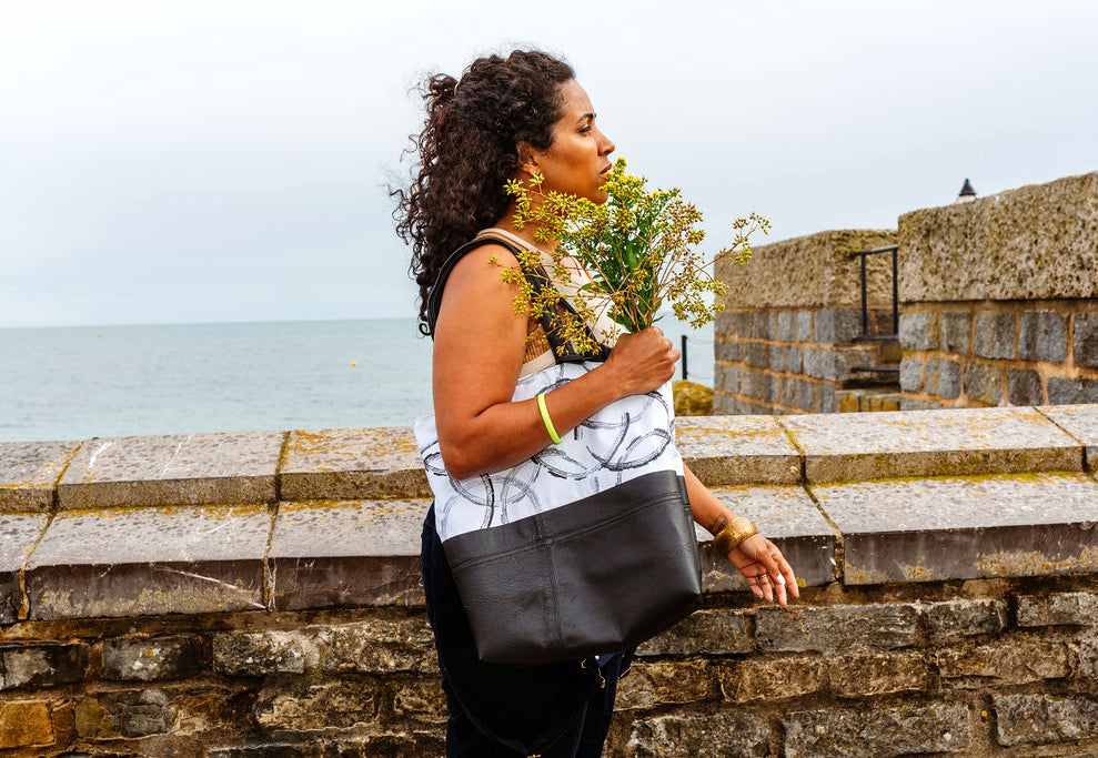 A black woman is carrying a hand printed leather and cotton tote bag, made from reclaimed materials. In her right hand she is holding yellow flowers. She is standing next to a sea wall and the sea is in the background.I